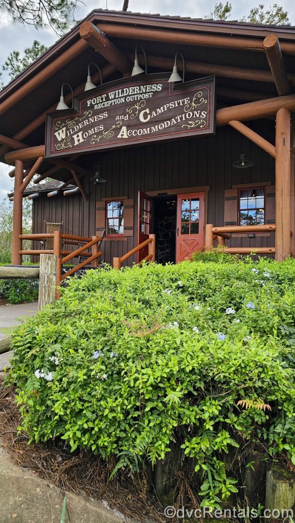 The log cabin-style exterior of the lobby of Disney’s Fort Wilderness Resort is seen under a cloudy sky. Above the doorway is a brown sign with white writing that reads “Fort Wilderness Reception Outpost. Wilderness Homes and Campsite Accommodations.”