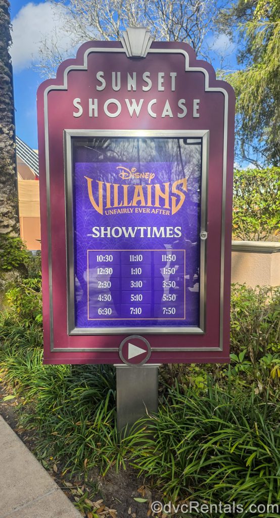 A purple and silver sign at Hollywood Studios’ Sunset Showcase displays showtimes for the Disney Villains: Unfairly Ever After show under a blue sky.