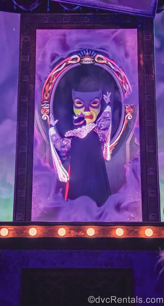 The Evil Queen interacts with the Magic Mirror on stage during a performance under hazy purple lighting effects.