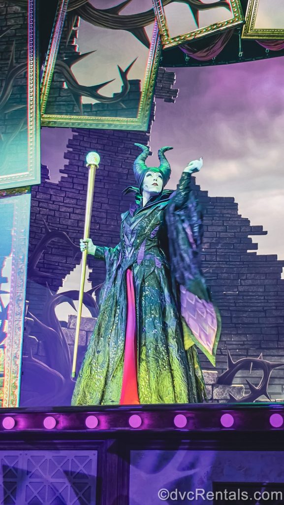 Maleficent poses with her horns held high as she performs on stage under purple and green lighting.