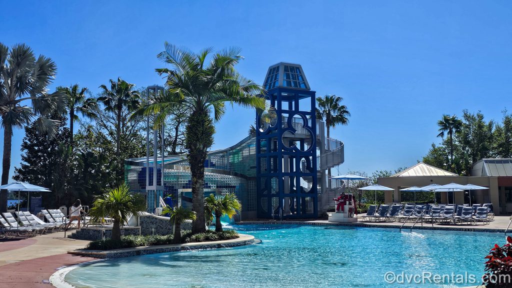 The blue waterslide tower featuring a Mickey-inspired design is seen in the distance with the Bay Cove Pool in the foreground under a bright blue sky at Bay Lake Tower.