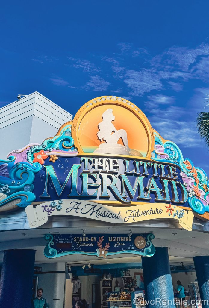 The attraction sign for The Little Mermaid – A Musical Adventure is seen over the entrance under a blue sky. The sign features a silhouette of Ariel in a golden circle, along with purple and blue nautical designs.