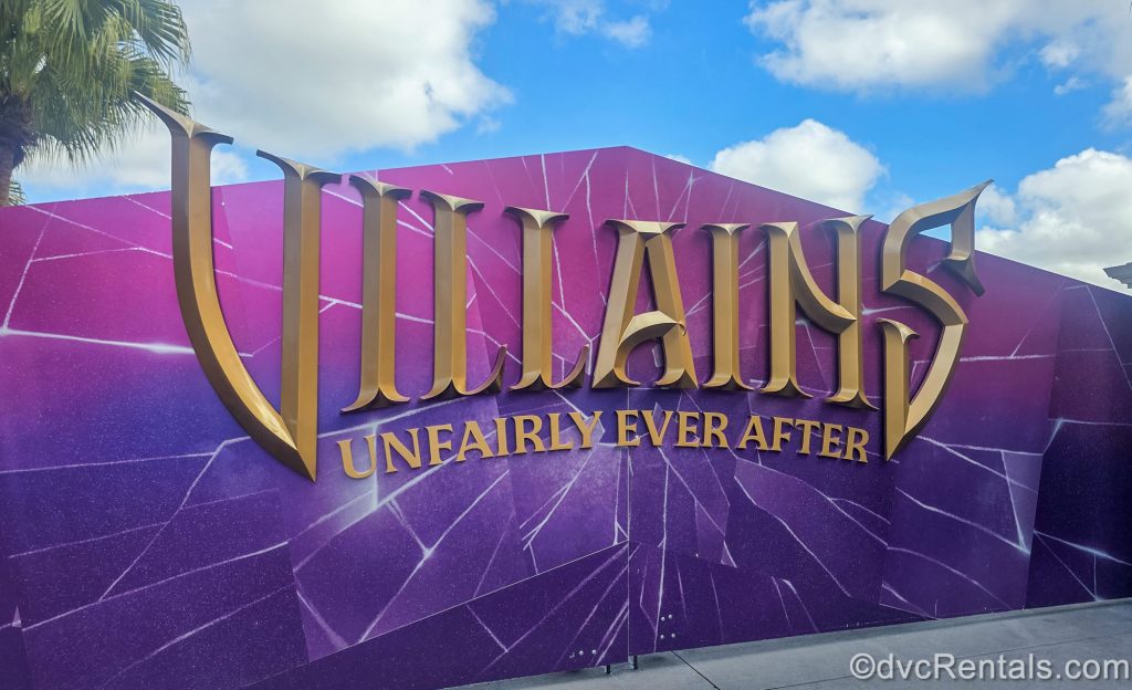 A large gate decorated pink and purple with the golden logo for “Villains Unfairly Ever After” is seen under a blue sky at Hollywood Studios.