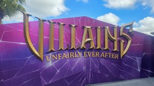 A large gate decorated pink and purple with the golden logo for “Villains Unfairly Ever After” is seen under a blue sky at Hollywood Studios.