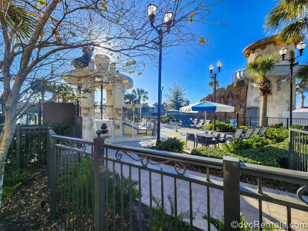 The entry way into the Riviera Pool is seen under the sunshine. Lounge chairs can be seen by the pool, along with the S’il Vous Play splash pad area.