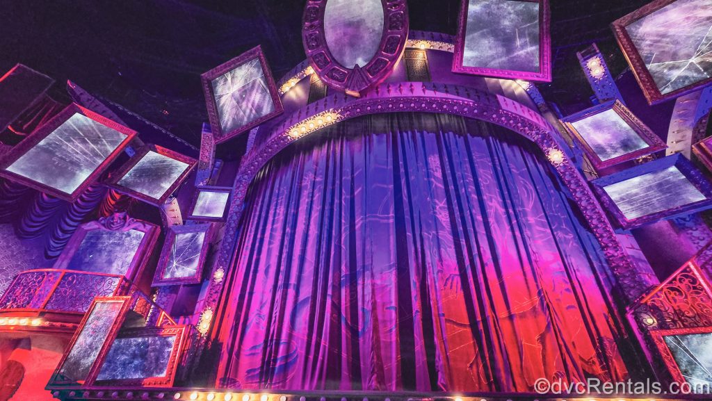 The stage is set for the Villains Unfairly Ever After show at Disney’s Hollywood Studios. Multiple large mirrors surround the set as the curtain and stage design are lit with red and purple lighting.