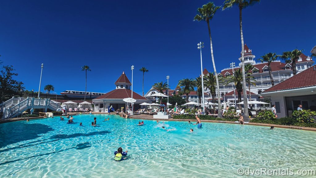 Guests enjoy a sunny day at the Beach Pool at Disney’s Grand Floridian Resort & Spa as the monorail glides past the resort in the background.