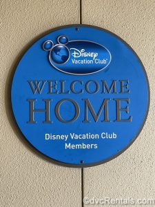 A circular blue sign reading “Disney Vacation Club. Welcome Home Disney Vacation Club Members” in white and silver lettering is posted on a beige wall.