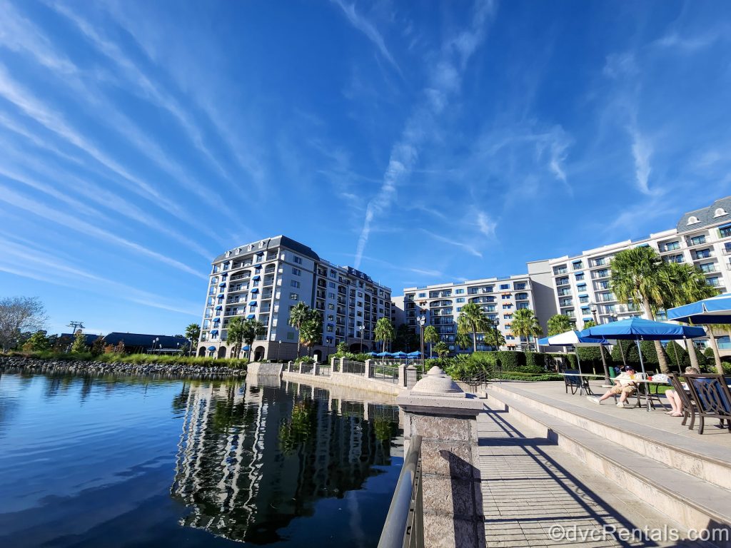 Guests sit at the umbrella-covered tables seen along the waterside at Disney’s Riviera Resort under a bright sunny sky.