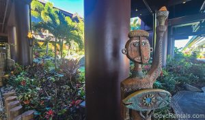 A carved Tiki figure holding a torch welcomes guests to Disney’s Polynesian Villas & Bungalows. Behind the statue the resort’s dark brown exterior as well as lots of tropical plants and flowers are seen.