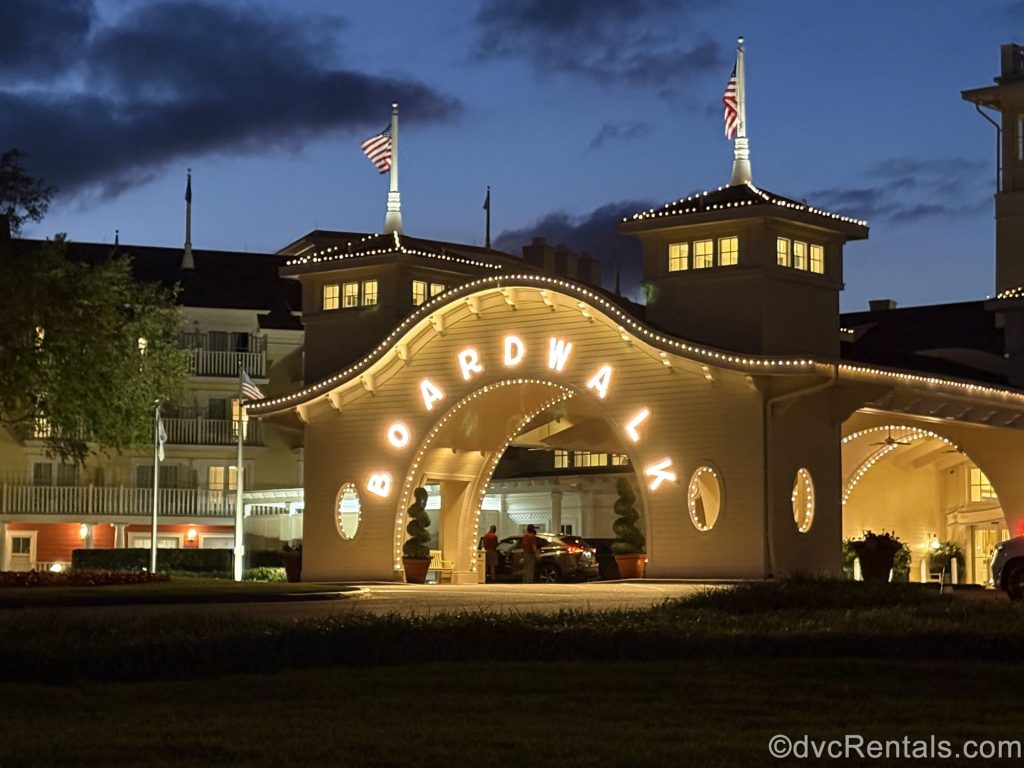 The word “BoardWalk” is spelled out in white lights over the main exterior entryway to Disney’s BoardWalk Villas under a hazy evening sky.