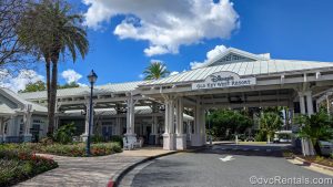 The drive-up entry way to Disney’s Old Key West Resort is seen under a blue sky. The building’s exterior is light blue and white and there are tropical flowers and palm trees in the surrounding landscaping.