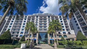 The white and grey exterior or Disney’s Riviera Resort is seen under a blue sky as palm trees line the pedestrian path leading to the building.