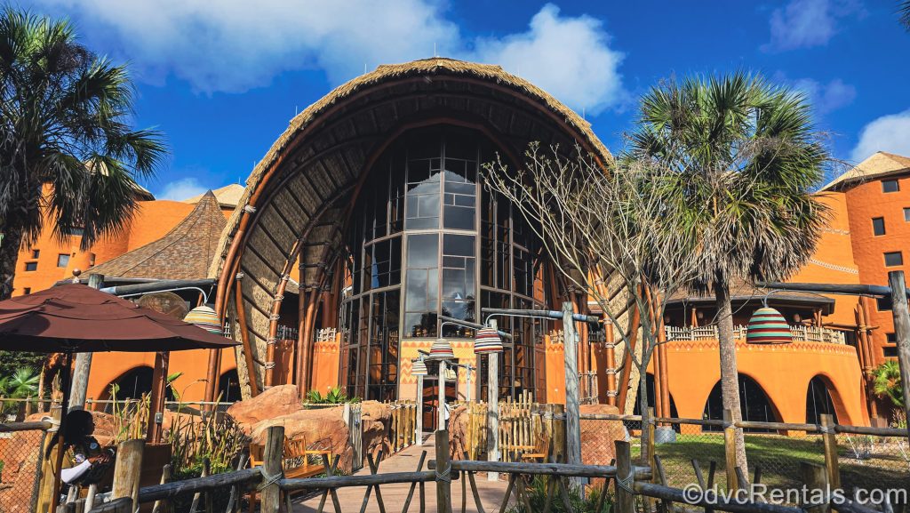 The exterior of Disney’s Animal Kingdom Villas Kidani Village is seen under a blue sky. The building is brown with multicolored accents inspired by African art.