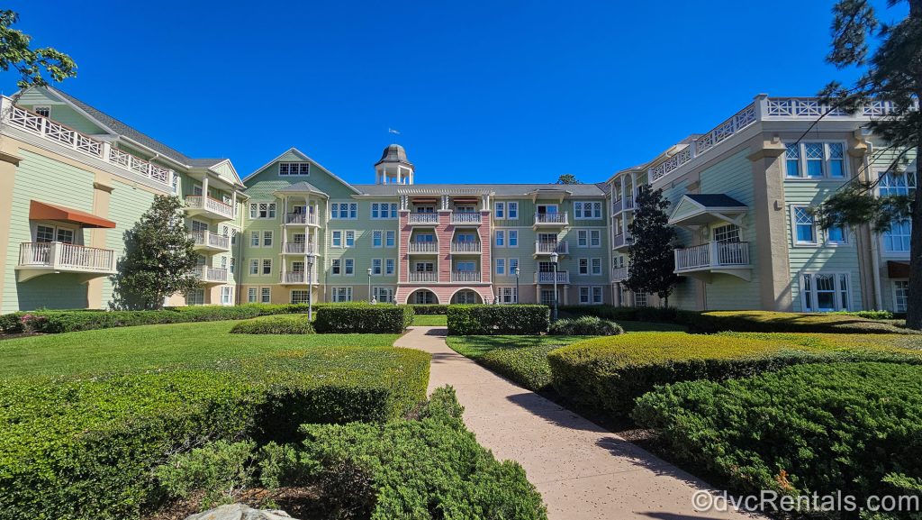 A pedestrian walkway and the pink, green, and yellow pastel exterior buildings of the resort are seen under a bright blue sky at Disney’s Saratoga Springs Resort & Spa.