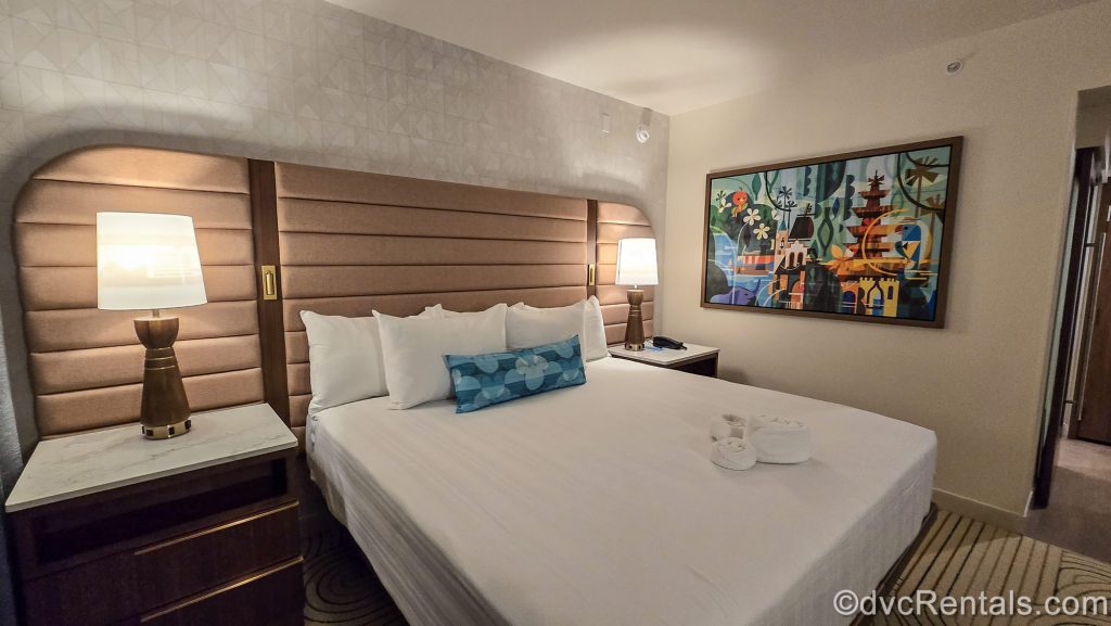 The king bed in the primary bedroom of a One Bedroom Villa at Bay Lake Tower at Disney’s Contemporary Resort. There is Mary Blair-inspired art on the wall and accent pillow, as well as a towel folded into a Mickey shape on the bed.