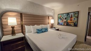 The king bed in the primary bedroom of a One Bedroom Villa at Bay Lake Tower at Disney’s Contemporary Resort. There is Mary Blair-inspired art on the wall and accent pillow, as well as a towel folded into a Mickey shape on the bed.