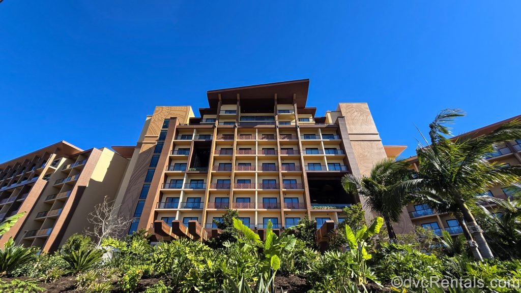 Lush green landscaping and the light and dark brown exterior of the Island Tower at Disney’s Polynesian Villas & Bungalows are seen under a bright blue sky.