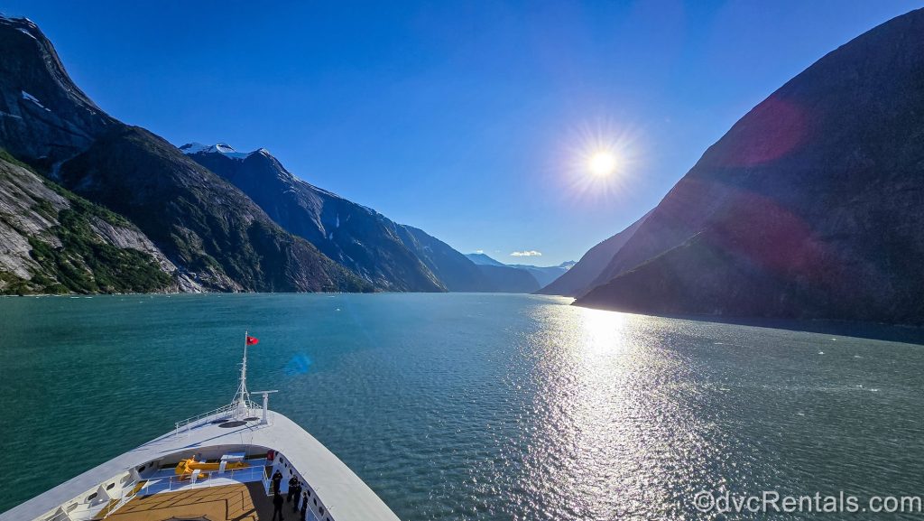 The Disney Magic sails through Endicott Arm Fjord in Alaska under a bright blue sky as mountains line the shore and fill the landscape in the distance.