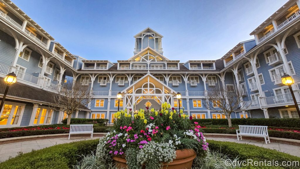The blue and white exterior of Disney’s Beach Club Villas is seen under a light blue sky as the sun begins to rise over the warm glow of the resort. In front of the entry way there are yellow, white, and pink flowers in the landscaping.