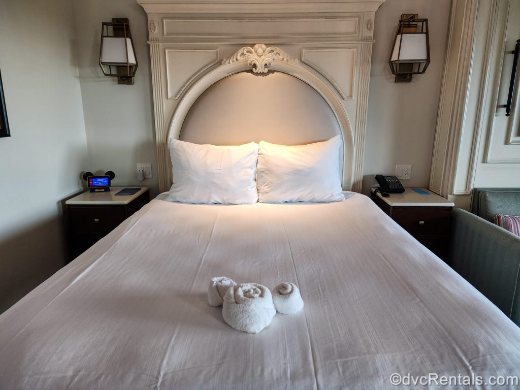 A queen bed in a Deluxe Studio at Disney’s Riviera Resort is seen. The bedding is white and there is a hidden Mickey made of white towels at the base of the bed.