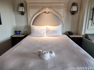A queen bed in a Deluxe Studio at Disney’s Riviera Resort is seen. The bedding is white and there is a hidden Mickey made of white towels at the base of the bed.