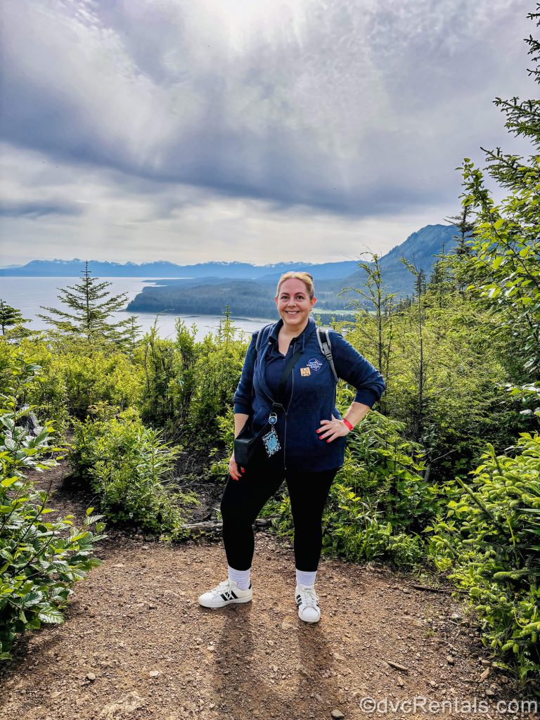 David’s Vacation Club Rentals team member Stacy R. poses along a hiking trail with the mountains and ocean in the distance.