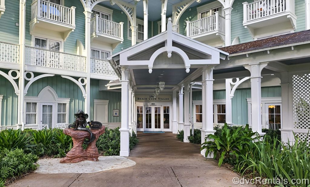 The pale green and white exterior of the DVC Villas building at Disney’s Beach Club Resort is seen. In front of the doors is a statue of Ariel from the Little Mermaid with a sign reading “Disney Vacation Club”.