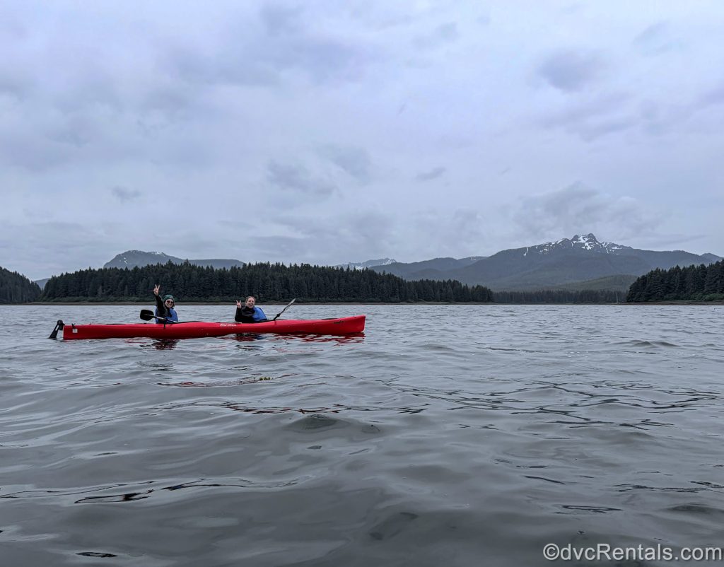 David’s Vacation Club Rentals team members Olivia and Stacy R. pose for a photo during their kayaking excursion in Icy Strait Point, Alaska, with mountains and wilderness in the background.