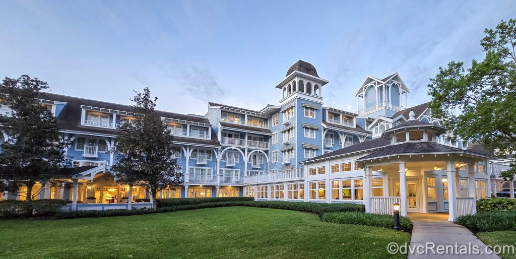 The light blue and white exterior of Disney’s Beach Club Resort is seen under a hazy morning sky as warm lights glow inside the resort.
