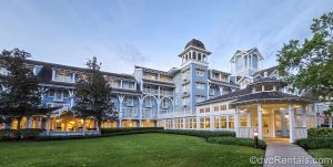 The light blue and white exterior of Disney’s Beach Club Resort is seen under a hazy morning sky as warm lights glow inside the resort.