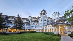 The light blue and white exterior of Disney’s Beach Club Resort is seen under a hazy morning sky as warm lights glow inside the resort.