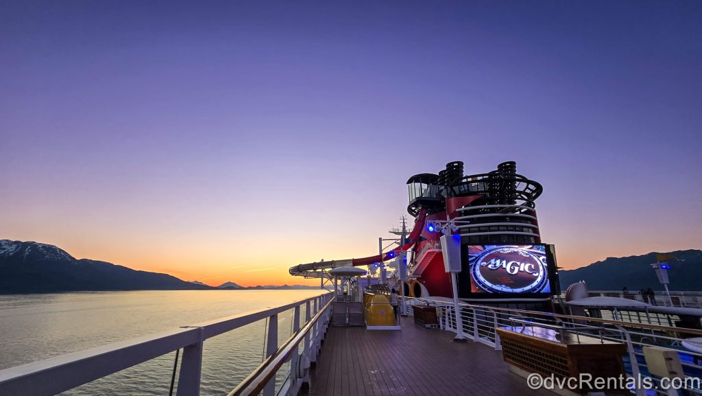 The Disney Magic sails through a mountainous Alaskan landscape as the sunset is overhead with tones of purple, orange, and yellow.