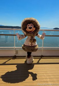 Minnie Mouse poses on the Disney Magic in her Alaskan coat and boots under a bright sun-filled sky with the ocean and mountains in the background.