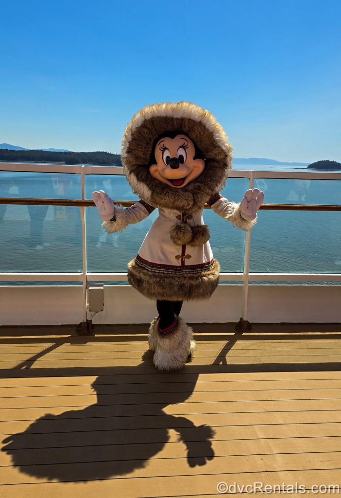 Minnie Mouse poses on the Disney Magic in her Alaskan coat and boots under a bright sun-filled sky with the ocean and mountains in the background.