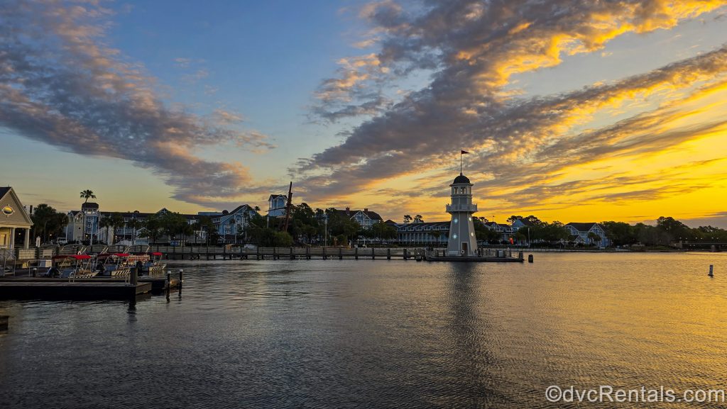 The exterior of Disney’s Beach Club Villas is seen under a beautiful yellow and blue sunrise sky across the water of Crescent Lake from Disney’s BoardWalk Villas.