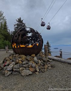 A large metallic ball-shaped fire pit with “Icy Strait Point” and a wilderness landscape carved into it holds a large bonfire as gondolas travel up to the mountain top behind under a cloud-filled sky.