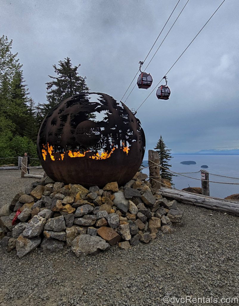 A large metallic ball-shaped fire pit with “Icy Strait Point” and a wilderness landscape carved into it holds a large bonfire as gondolas travel up to the mountain top behind under a cloud-filled sky.