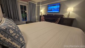 A Deluxe Studio at Disney’s Beach Club Villas. The bed is decorated with white linen and a blue pillow with Daisy Duck, and looks towards the room’s television and patio door.