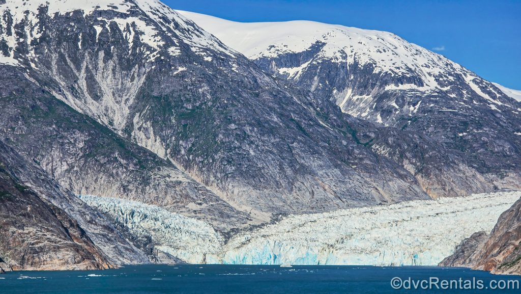 The white and blue ice of Dawe’s Glacier is seen nestled in the mountains against the blue waters of the Endicott Arm Fjord under a clear blue sky.