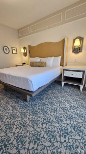 The king-sized bed in the One Bedroom room size at Disney’s BoardWalk Villas. There is white bedding with themed accents and patterned carpet with a blue print.