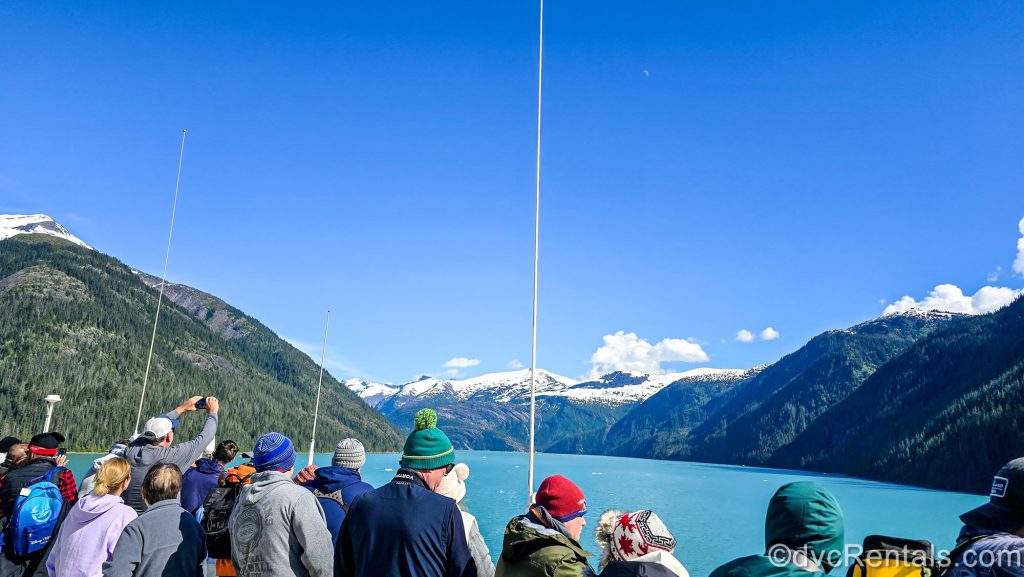 Cruise guests on the Disney Magic enjoy the amazing view of the mountainous Alaskan landscape from the upper outdoor decks as the ship sails through the Endicott Arm under a blue sky.
