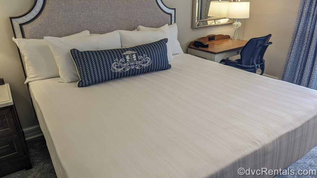 white king bed with a grey fabric headboard and blue pillow featuring Donald and Daisy Duck is seen next to a small wooden desk in a Two Bedroom Villa at Disney’s Beach Club Villas.
