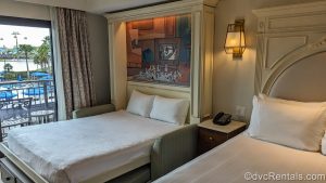 The queen size bed and pull-down Murphy bed in the Deluxe Studio at Disney’s Riviera Resort. The resort grounds and Disney Skyliner are visible outside the balcony door, and there is a 101 Dalmatians-theme mural behind the Murphy bed.