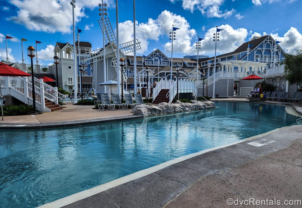 A portion of the lazy river in the Stormalong Bay pool complex is seen under a blue sky. The resort exterior and nearby lounge chairs are also seen as guests relax by the water.