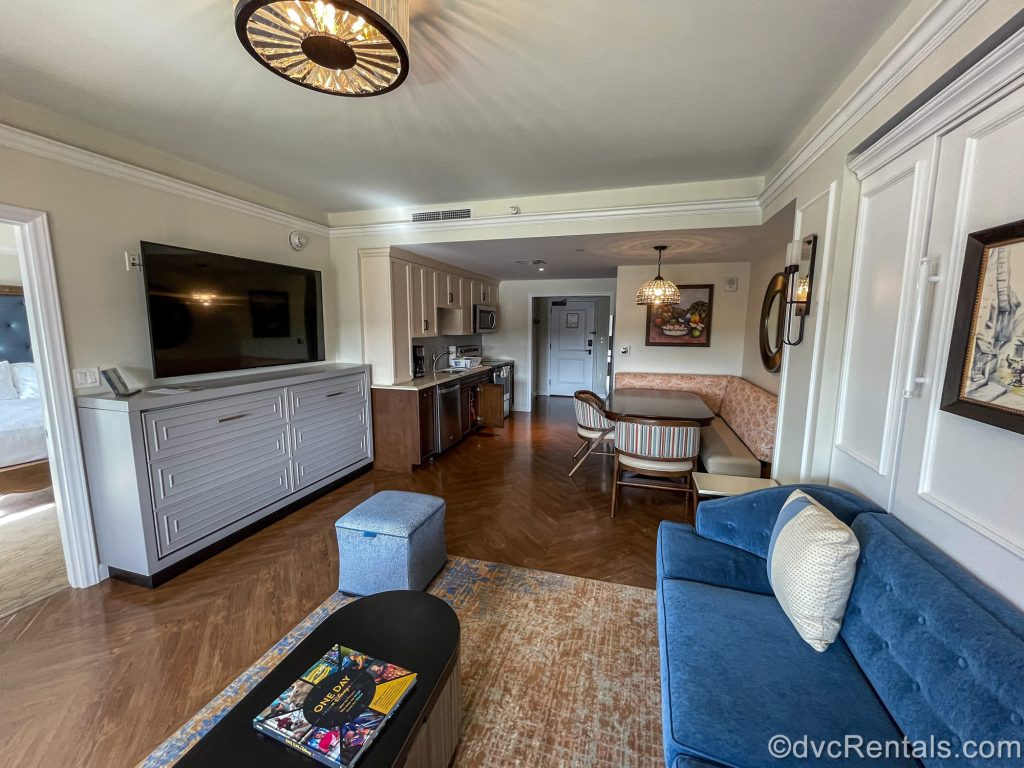 The living room, dining area, and kitchen in a One Bedroom Villa at Disney’s Riviera Resort. The room is decorated with pops of gemstone colors and Disney artwork inspired by classic European works.