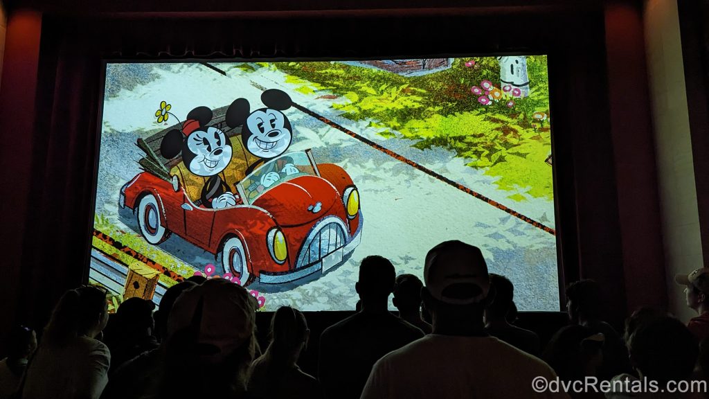 Park goers watch as Mickey and Minnie Mouse are seen driving in a little red convertible in an animated video shown on screen during the pre-show segment of Mickey & Minnie’s Runaway Railway.
