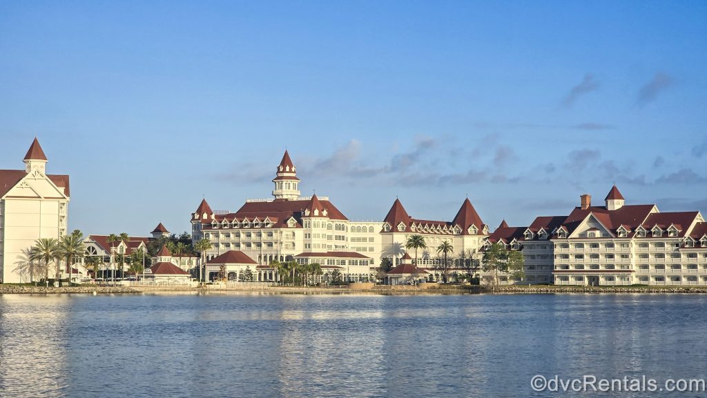 The white and red exterior of Disney’s Grand Floridian Resort & Spa is seen from across the Seven Seas Lagoon under a blue sky.