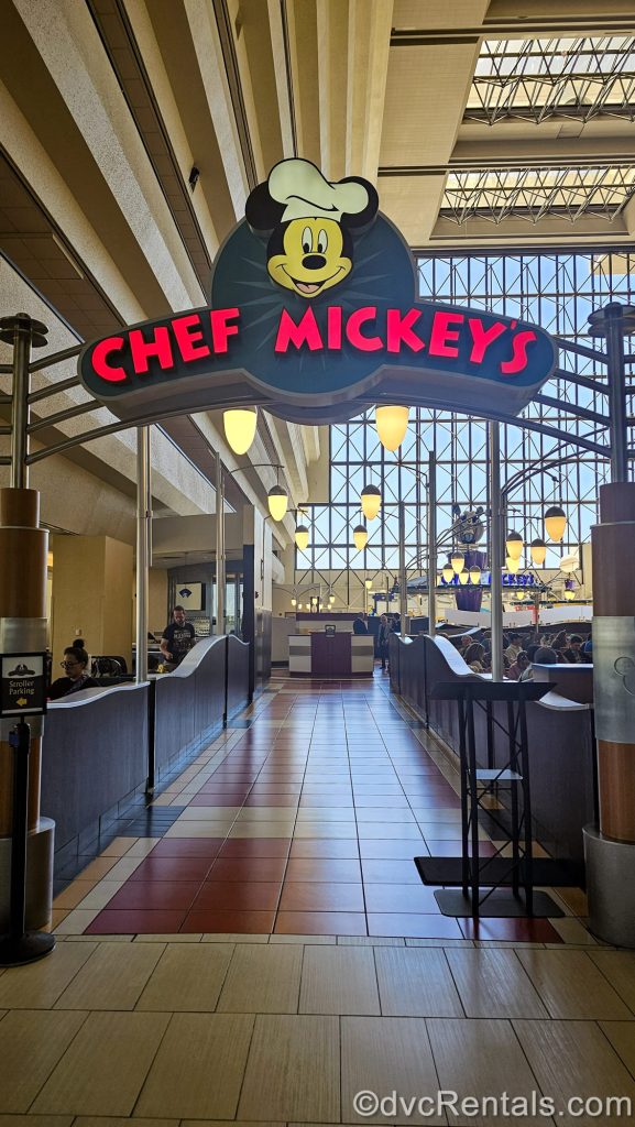 Mickey Mouse is seen in a chef’s hat atop the sign for Chef Mickey’s at Disney’s Contemporary Resort. The restaurant’s seating area and resort’s tall glass windows are visible in the background.