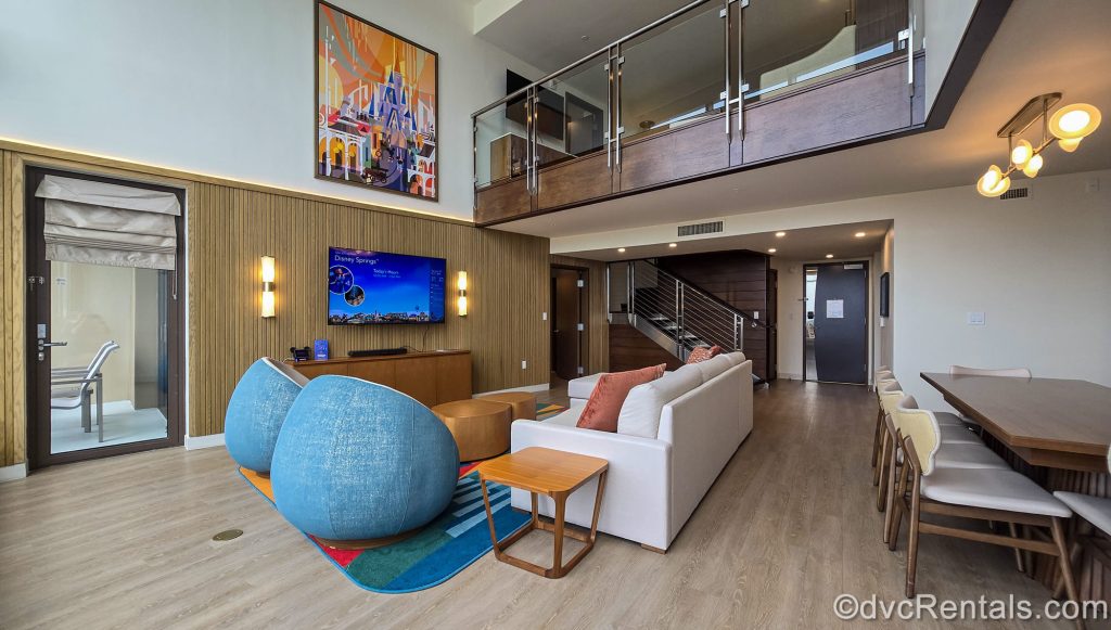 The living room and a portion of the dining area of a Three Bedroom Grand Villa at Bay Lake Tower at Disney’s Contemporary Resort. The room is decorated with wooden tones and pops of bright colors, along with Mary Blair-inspired art.